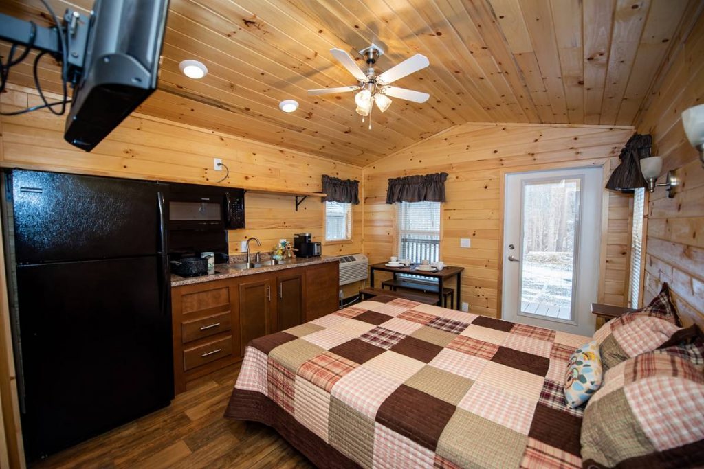 Boo Boo Two Room Cabin interior, kitchen and living area with TV, refrigerator, microwave, stovetop, ceiling fan, dining table and dishes, and queen-size bed