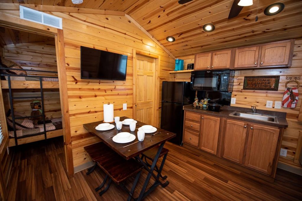 Cindy bear cabin interior, dining area with dining table, dishes, TV, microwave, refrigerator, stovetop, cabinets, and sink