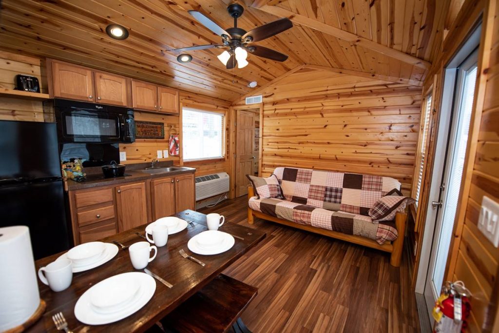 Cindy Bear cabin interior, kitchen and living space with full-size futon, dining table with dishes, refrigerator, stovetop, microwave, sink, and ceiling fan