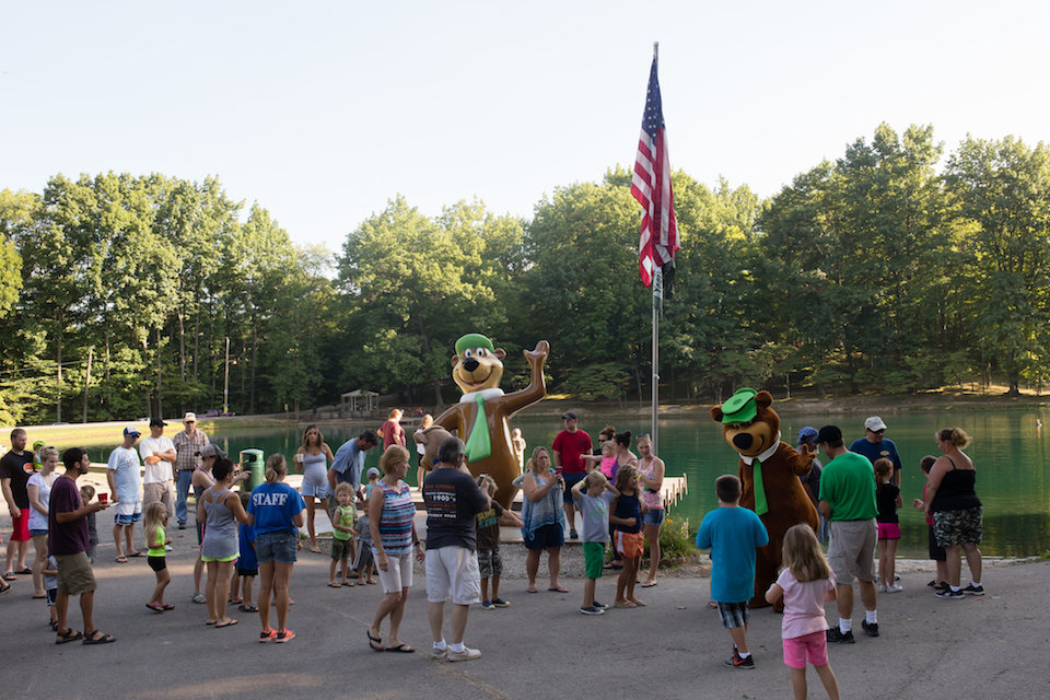 A crowd of campers and children surround a bear mascot, with an American flag and the lake behind them